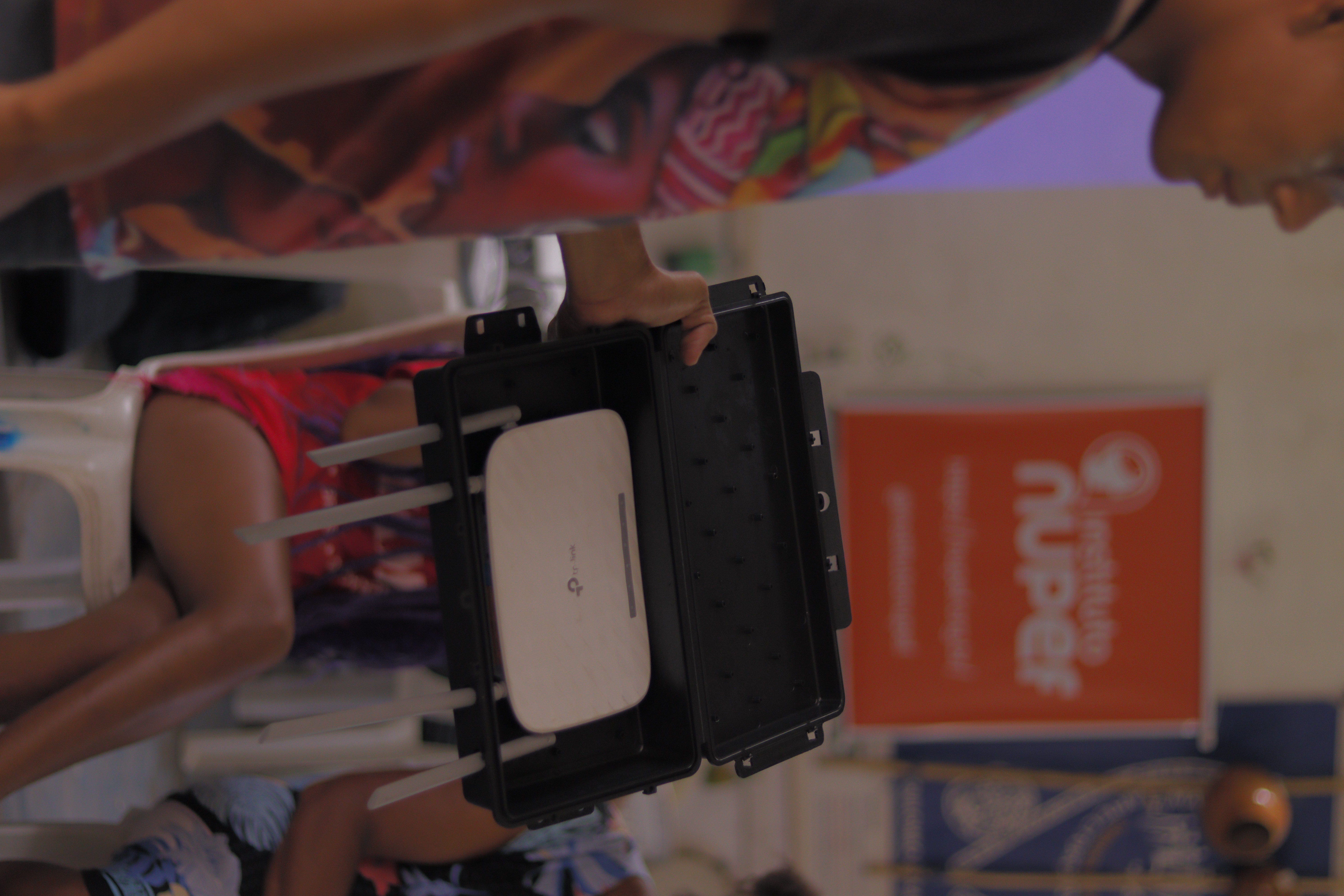 Pessoa em primeiro plano segura uma caixa plástica preta aberta contendo um roteador Wi-Fi branco com quatro antenas. Ao fundo, há pessoas sentadas em um ambiente de oficina ou apresentação, e um banner desfocado com o texto Instituto Nuper.