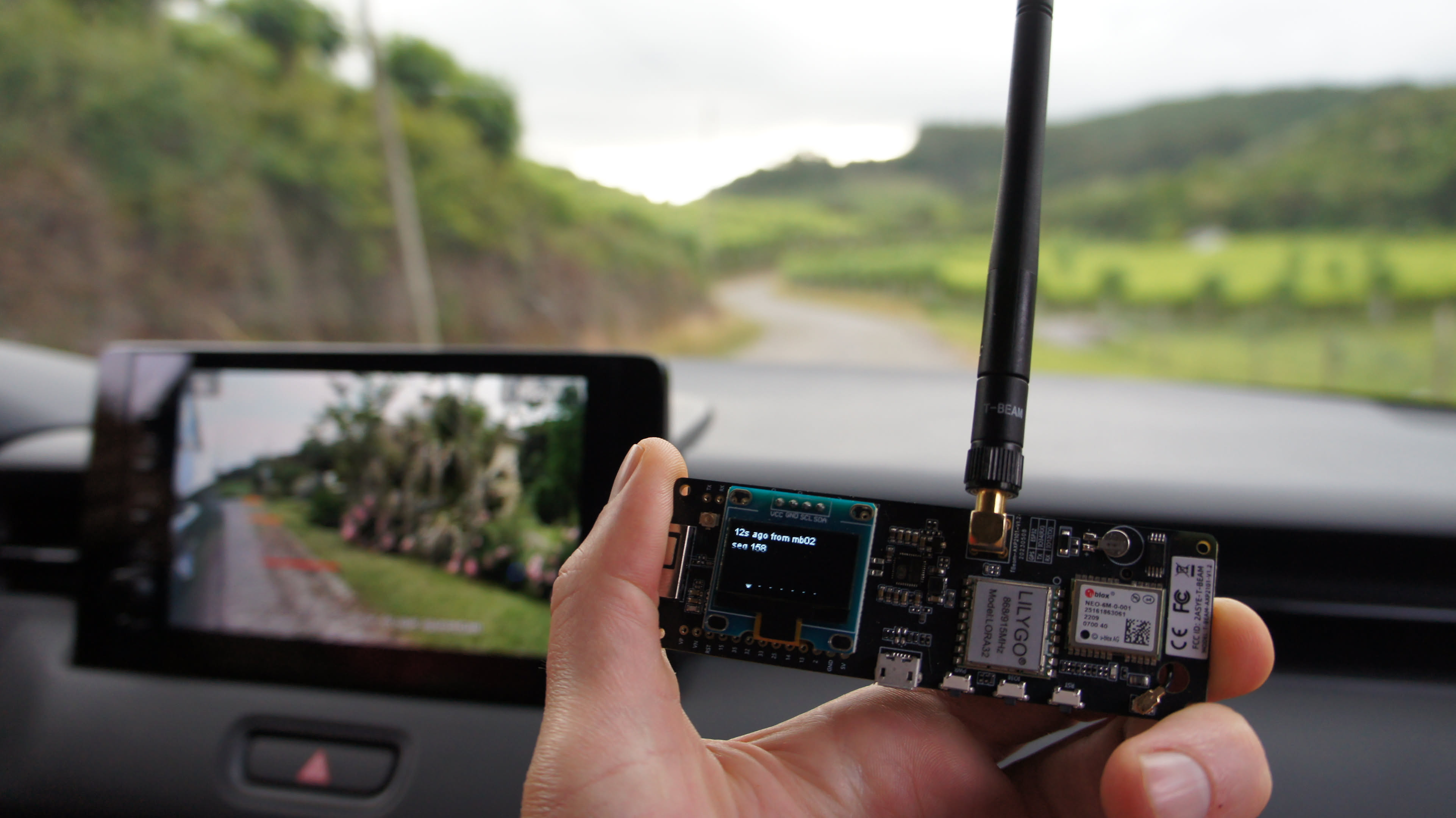 Mão segura um dispositivo LILYGO T-Beam com antena e tela OLED dentro de um carro em movimento, mostrando uma mensagem recebida via Meshtastic. Ao fundo, um celular preso ao painel exibe a estrada e a paisagem verde através da câmera.