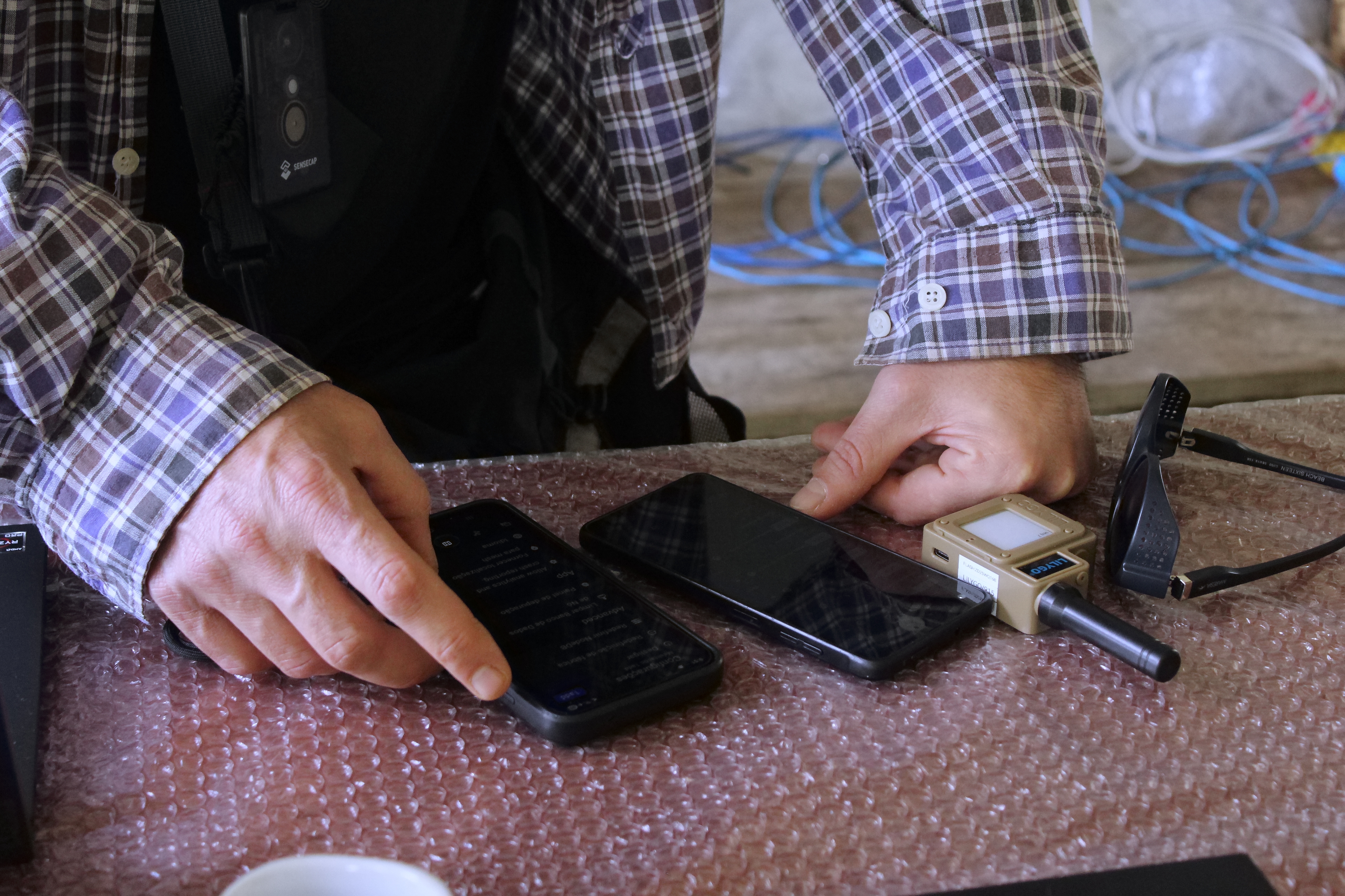Duas mãos manuseiam dois celulares sobre uma superfície com plástico bolha, ao lado de um dispositivo LILYGO com antena e um óculos de sol.