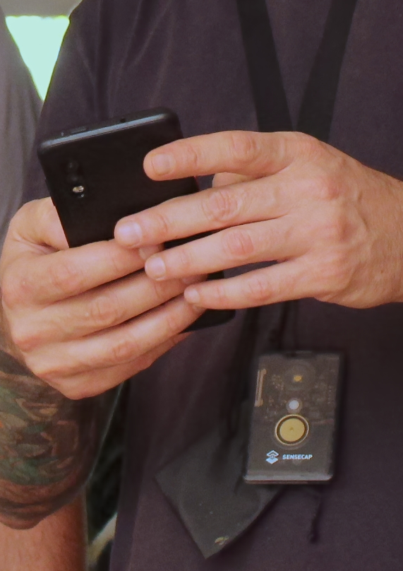 Close das mãos de uma pessoa segurando um celular preto, com um dispositivo SenseCap pendurado no pescoço por um cordão.