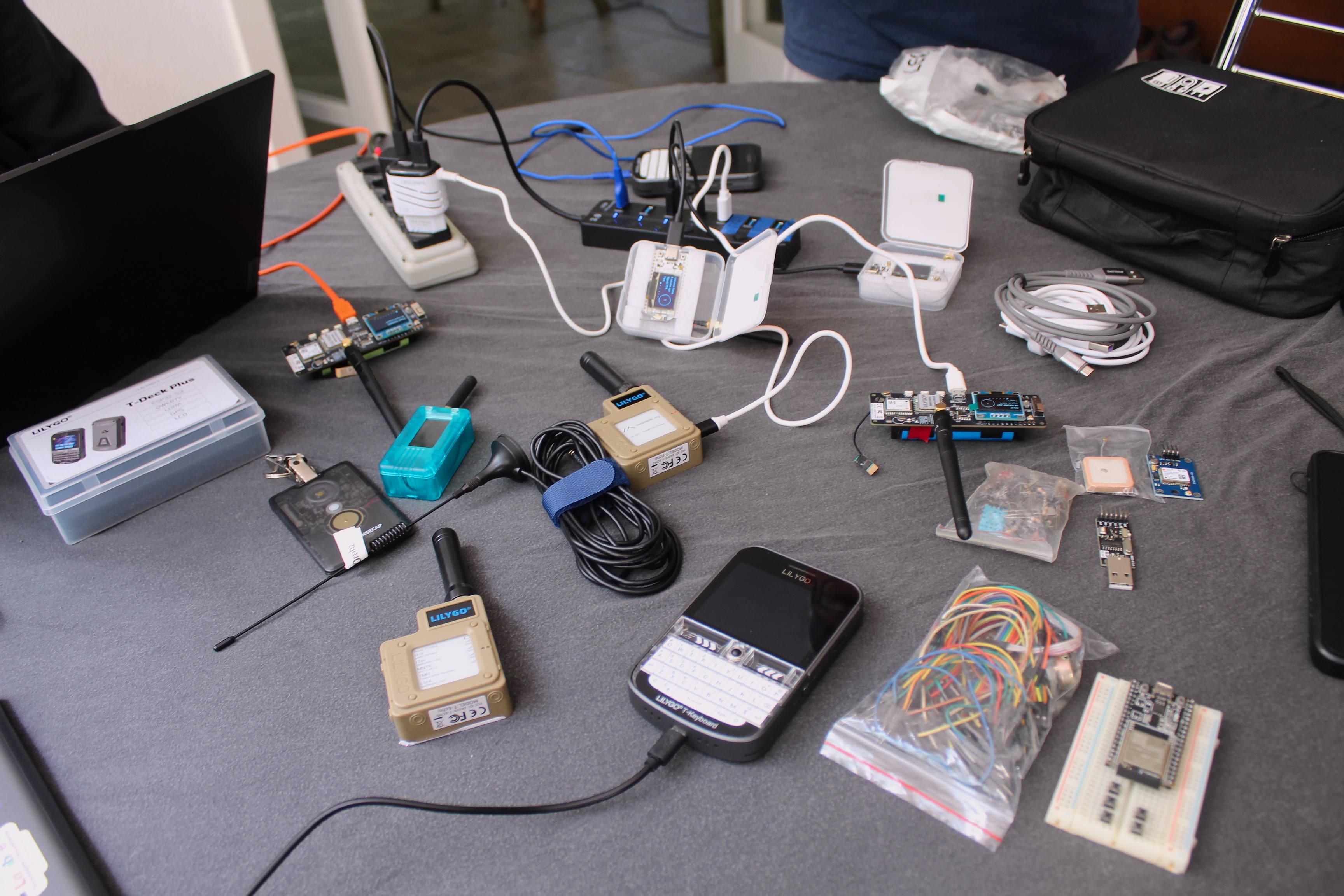Mesa com vários dispositivos Meshtastic e componentes eletrônicos organizados: nós LoRa com antenas, um T-Deck Plus, um T-Keyboard, cabos, uma protoboard, módulos GPS e sacos com componentes soltos.
