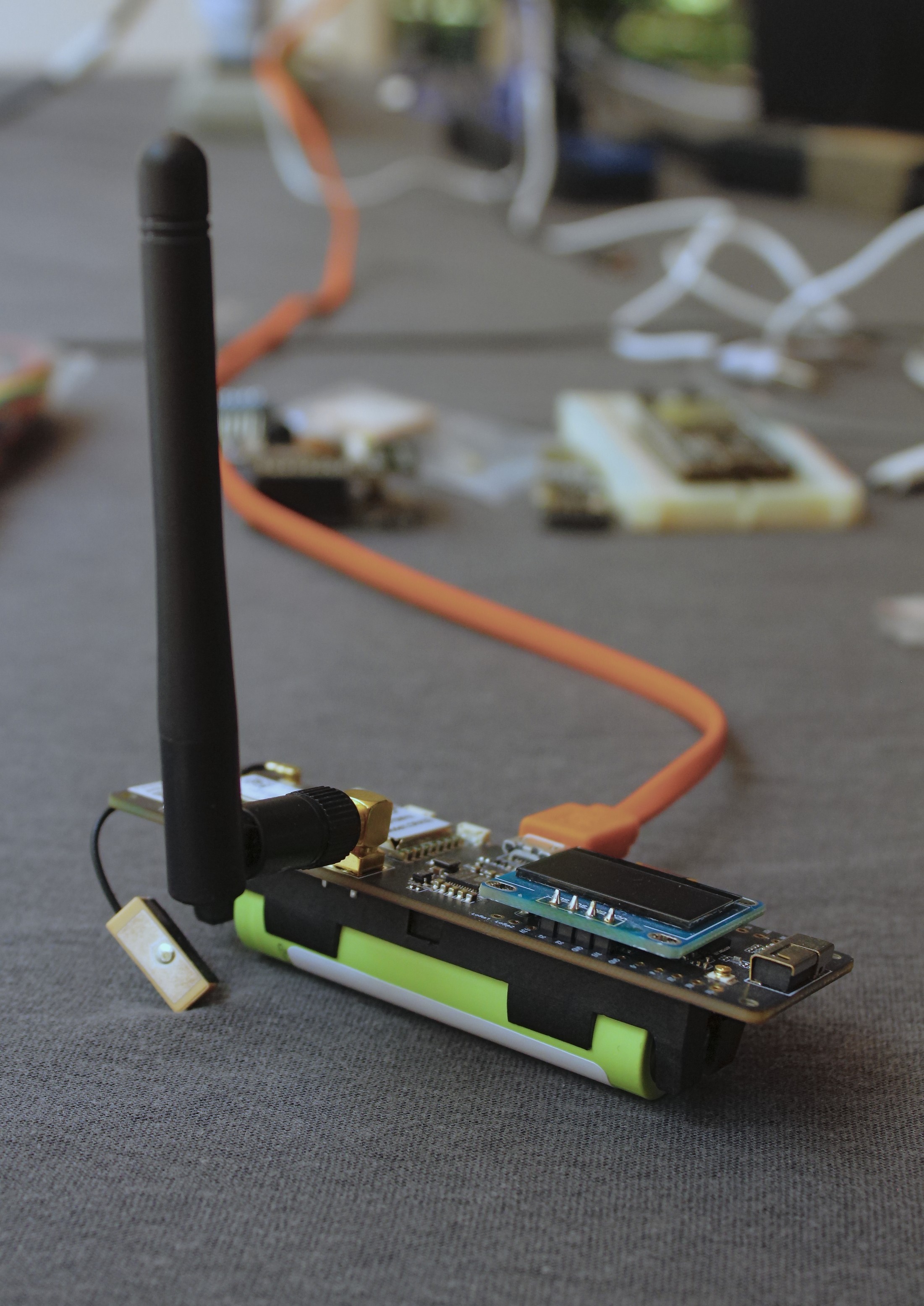 Nó Meshtastic com antena e bateria verde, tela OLED e módulo GPS conectado por um cabo curto, apoiado sobre um tecido cinza, com um cabo laranja ligado à porta USB.