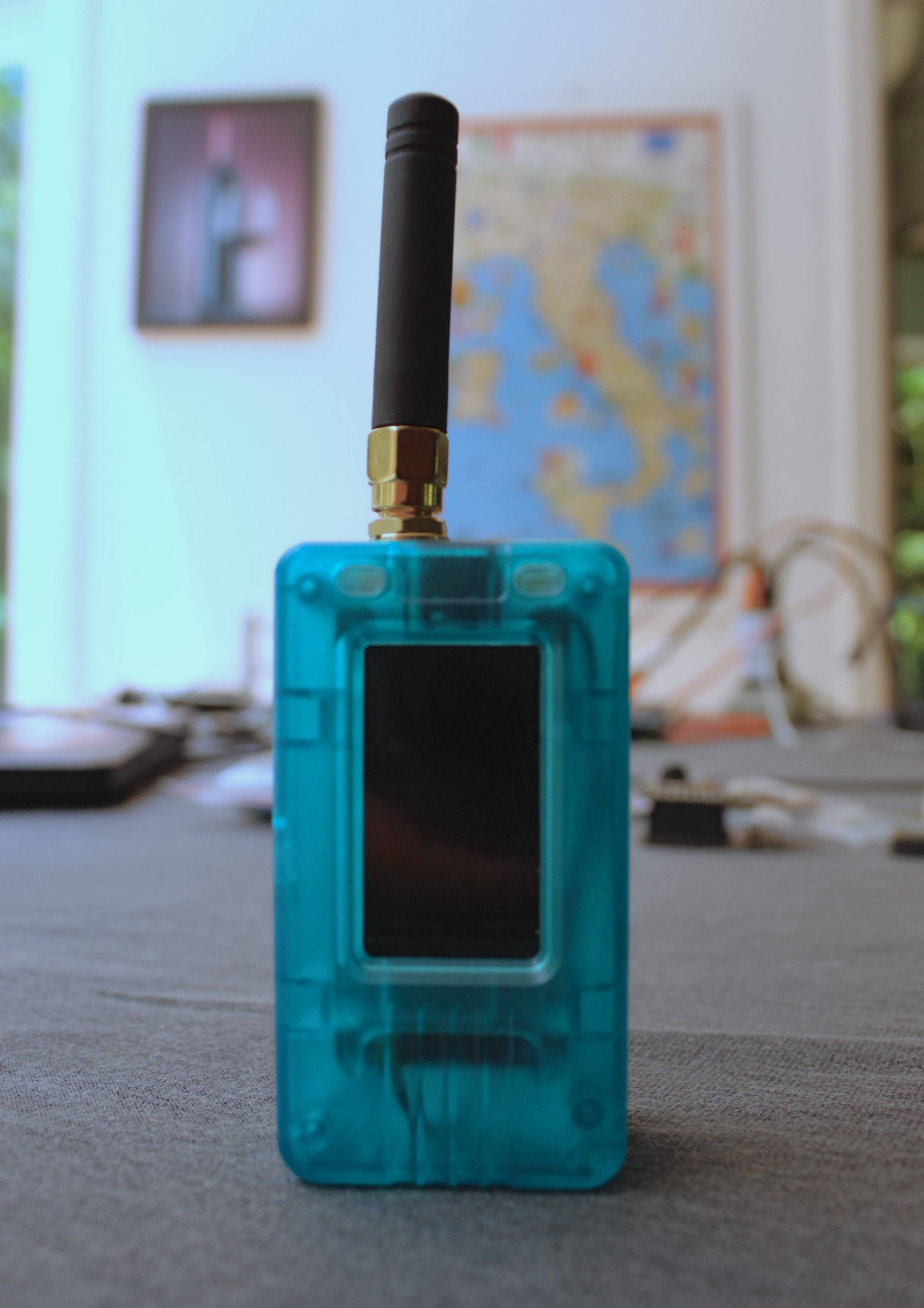 Dispositivo Meshtastic em case transparente azul turquesa com antena preta, em pé sobre uma mesa.