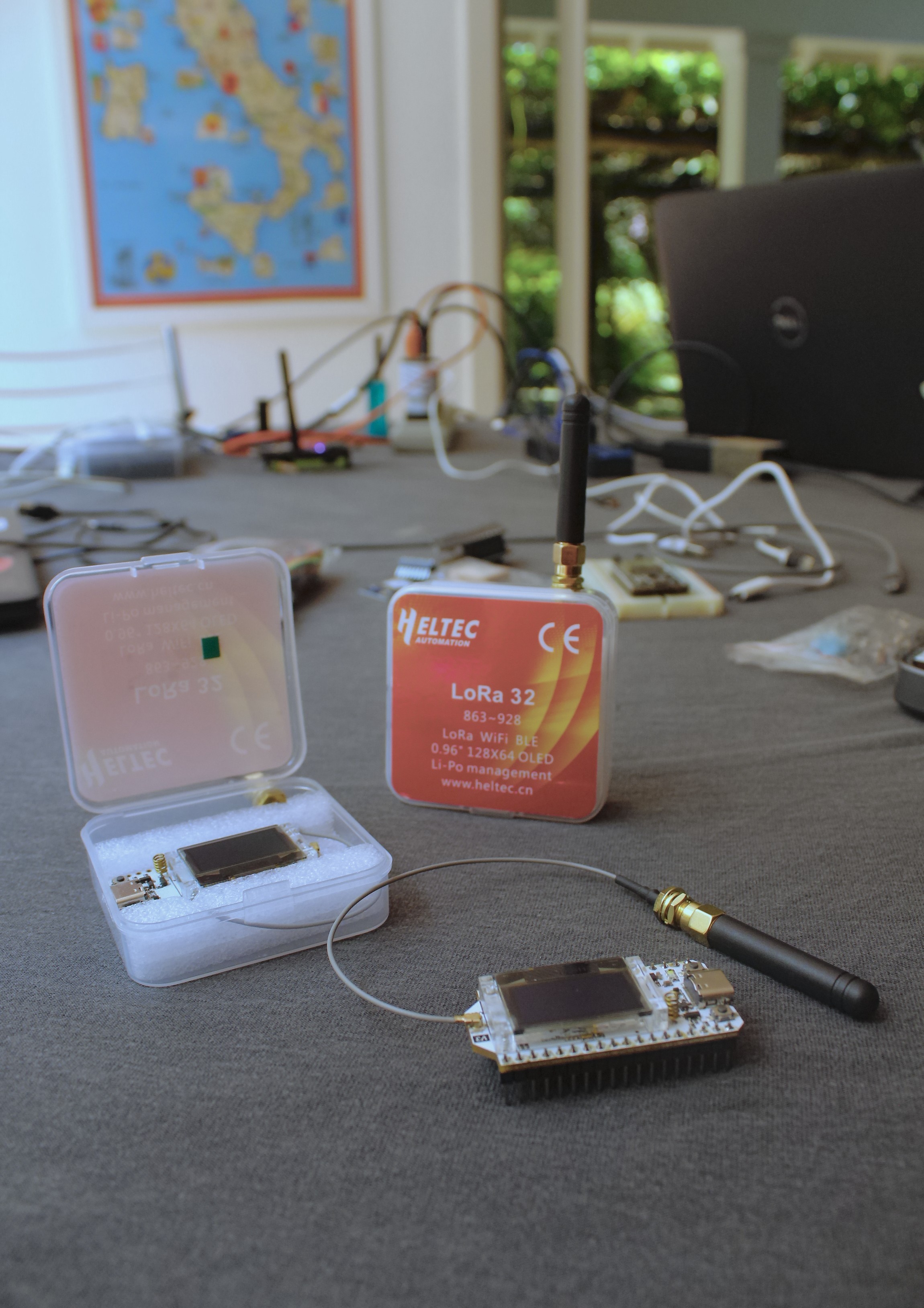 Dois dispositivos Heltec LoRa 32 sobre uma mesa, um dentro de uma caixa plástica com espuma e outro com antena conectada por cabo a uma placa com tela OLED.
