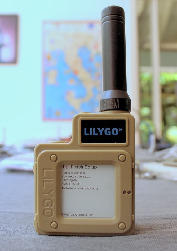 Dispositivo LILYGO bege com antena, exibindo na tela instruções de configuração inicial do Meshtastic (conectar antena, conectar aplicativo, definir região e fuso horário).