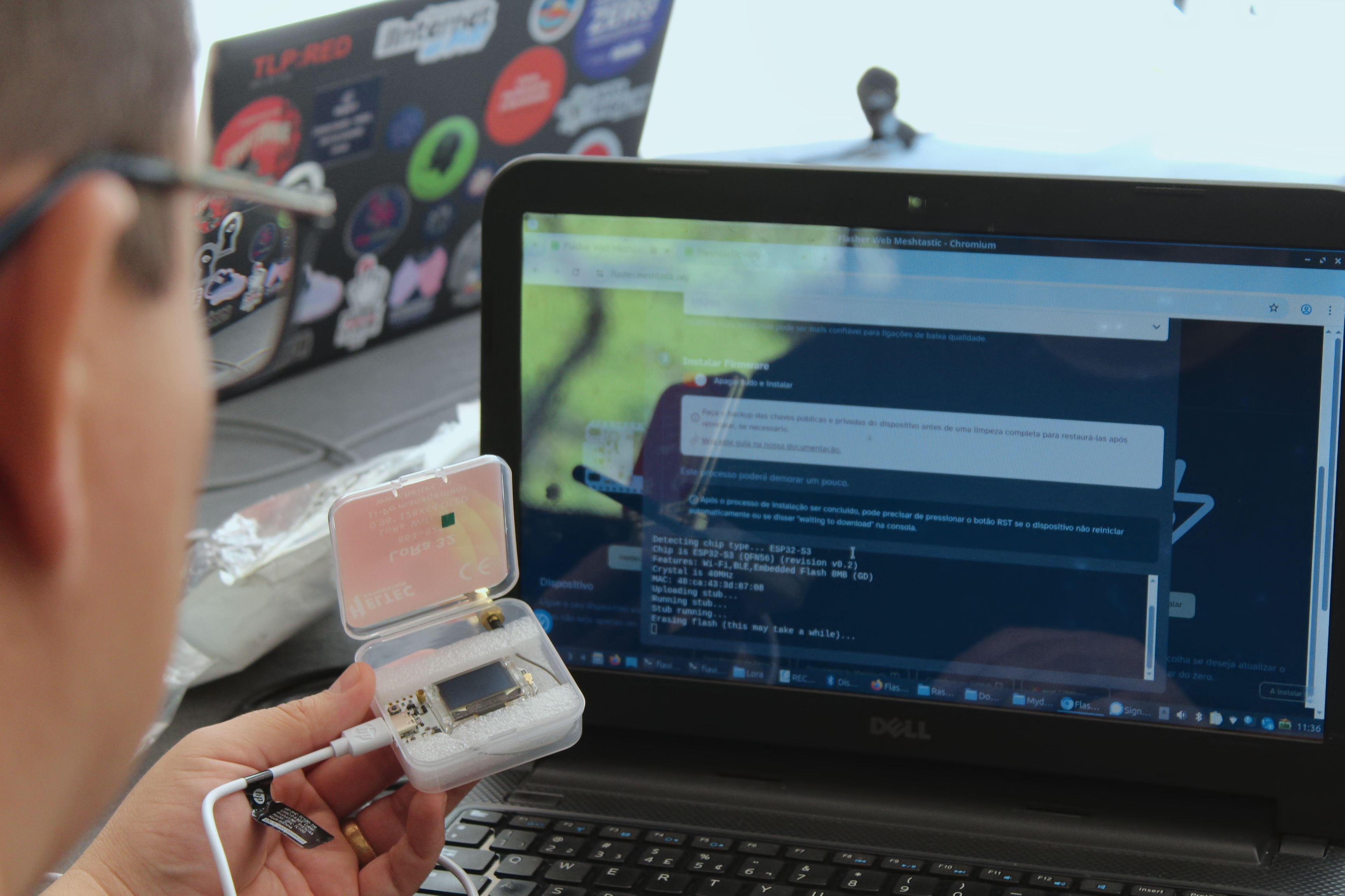 Pessoa de óculos segura um nó Meshtastic aberto dentro de uma caixa plástica com espuma protetora, enquanto grava o firmware por um notebook Dell que exibe o instalador web do Meshtastic em português.