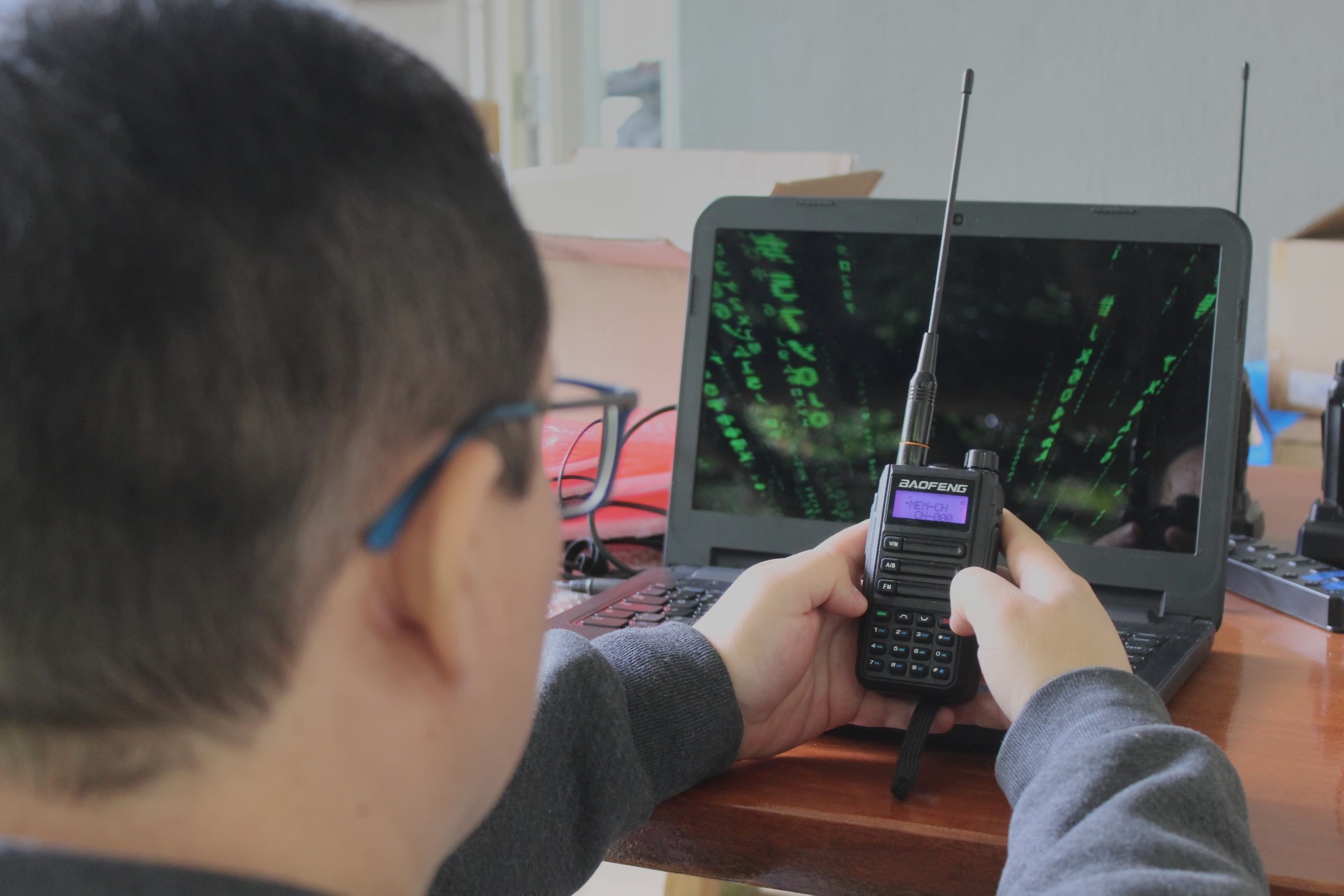 Pessoa de óculos segura um rádio portátil Baofeng com antena, testando a configuração de canal junto a um notebook que exibe uma tela de código em estilo matrix.