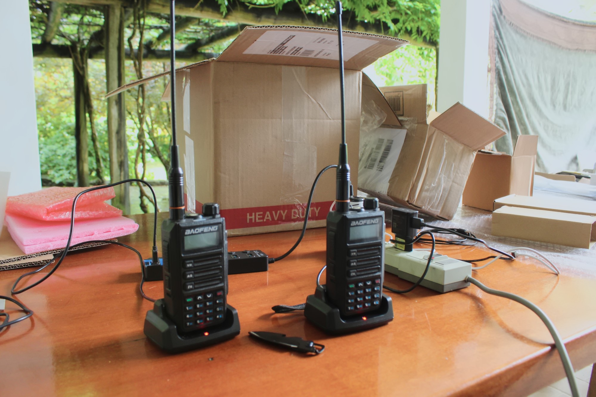 Dois rádios portáteis Baofeng com antenas, encaixados em bases carregadoras sobre uma mesa, com caixas de papelão ao fundo.