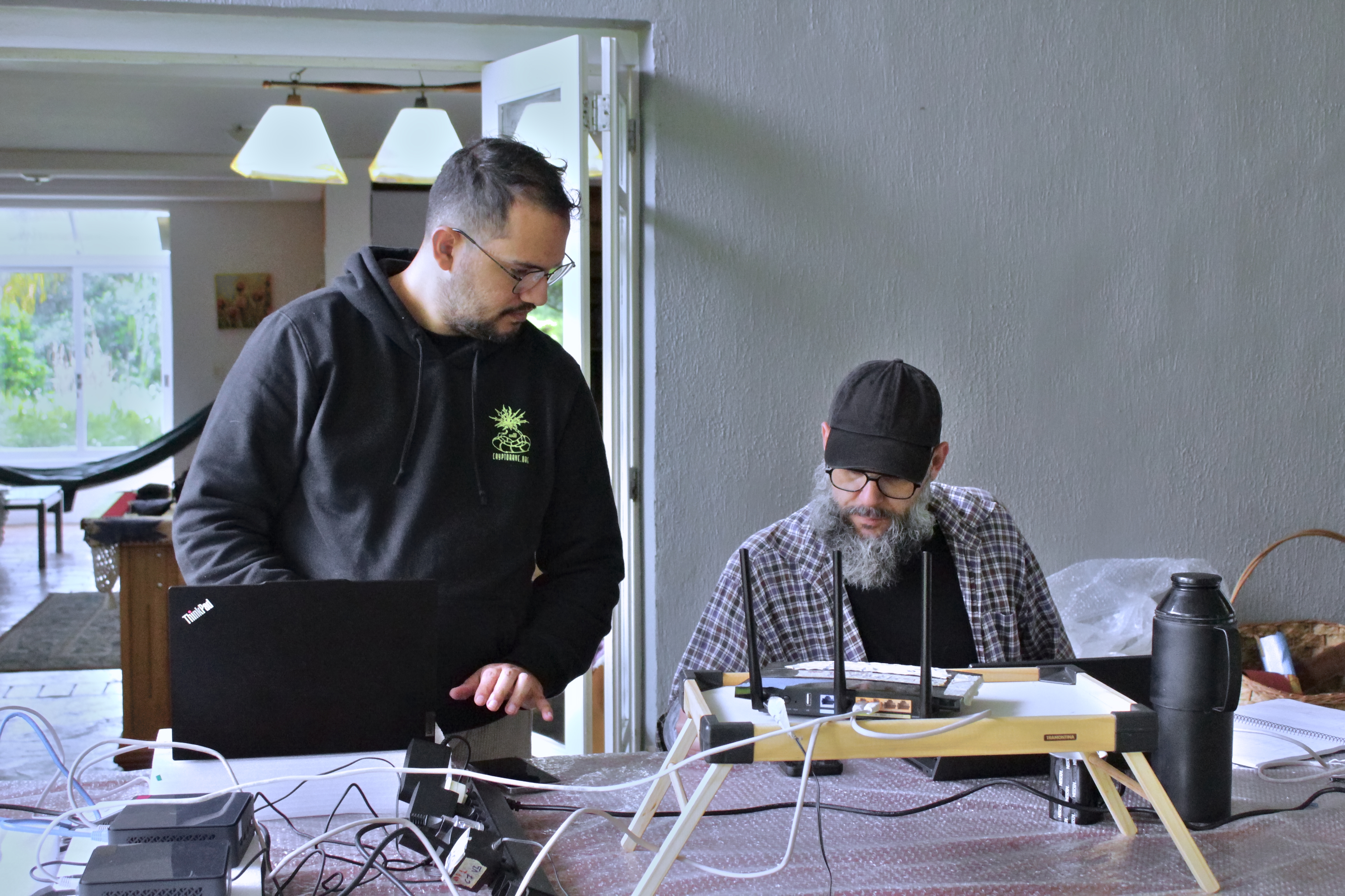 Duas pessoas trabalham em uma mesa com equipamentos de informática. À esquerda, uma pessoa em pé usa um notebook conectado por vários cabos. À direita, outra pessoa sentada observa um equipamento sobre um suporte, ao lado de um roteador com antenas e uma garrafa térmica preta. O ambiente é interno, com uma porta aberta e luz natural entrando pela lateral.
